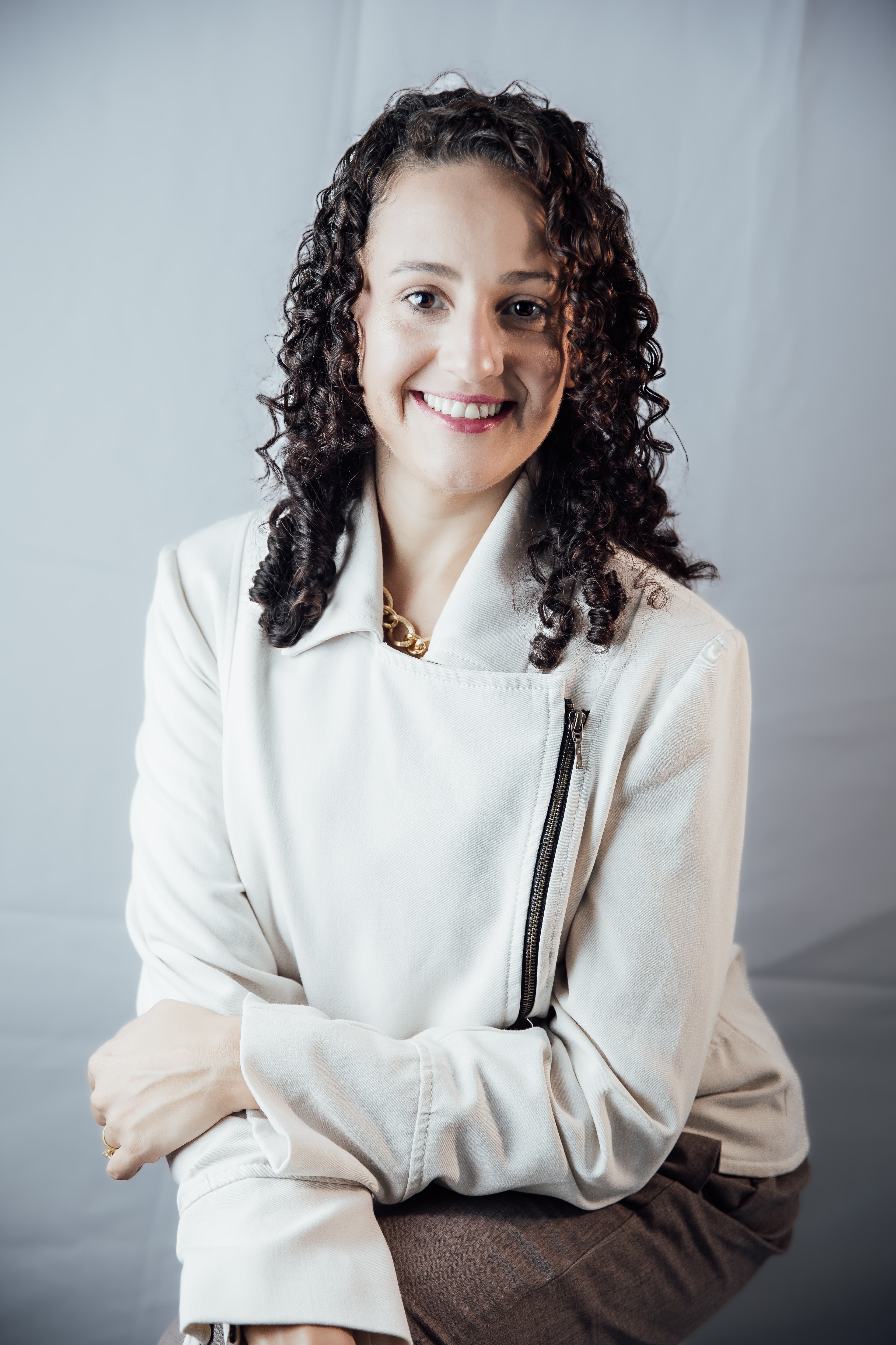Eloá Fraga sorrindo, de cabelo cacheado e jaqueta off-white, em retrato de estúdio com fundo claro